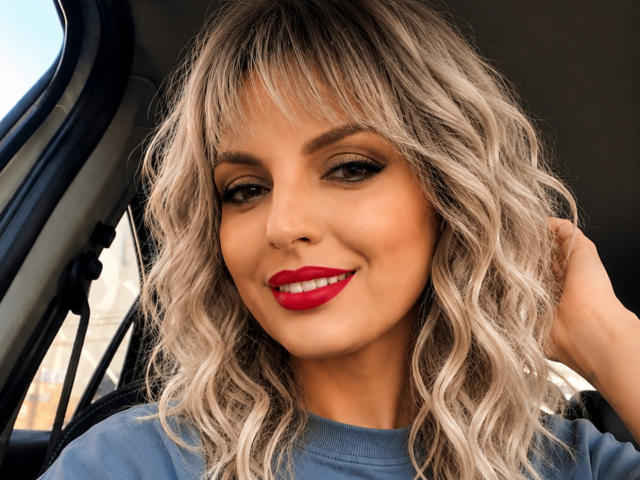 DouceAmelia with wavy blonde hair, red lipstick, and blue shirt, smiling in a car, hand in hair, looking cute with sexy makeup, showing her tits subtly.