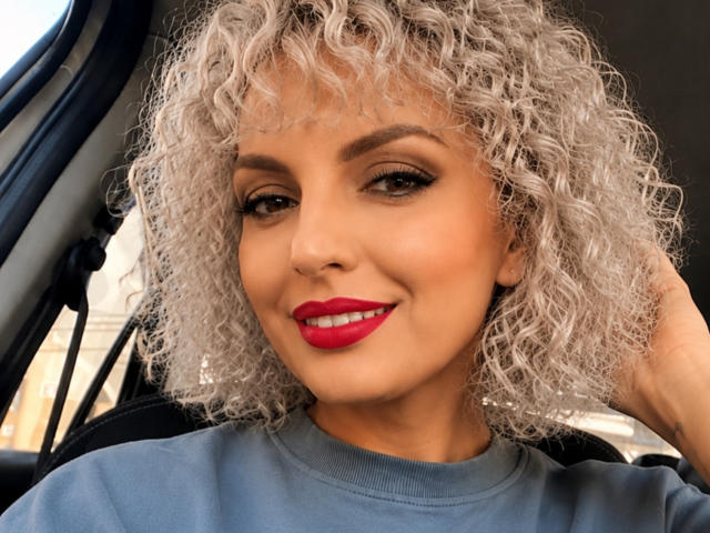 DouceAmelia, con i capelli grigi ricci, il rossetto rosso e una felpa grigia, sorride in macchina, con una mano che le tocca la testa. Ha un viso carino, sexy e attraente..