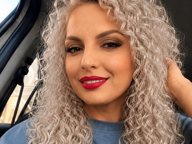 DouceAmelia ha i capelli grigi e ricci, il rossetto rosso e un sorriso, è seduta in macchina, indossa una camicia blu e ha una mano tra i capelli. Ha un viso carino..