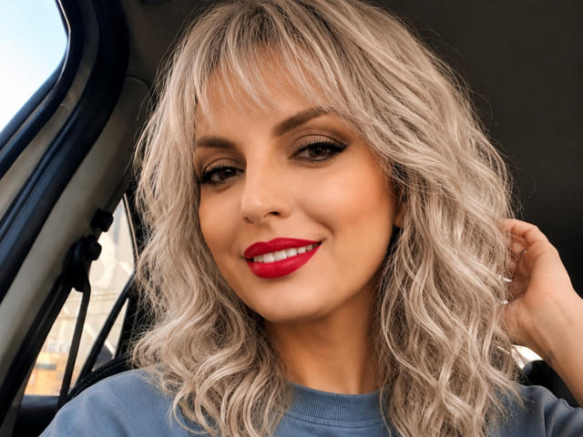 DouceAmelia, con i capelli biondi mossi, rossetto rosso e eyeliner scuro, sorride in macchina, indossa una camicia blu e si tocca i capelli..