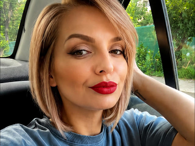 DouceAmelia has blonde bob, red lipstick, and blue eyes, with a tattoo on her left arm, wearing a blue shirt, sitting in a car, smiling subtly.