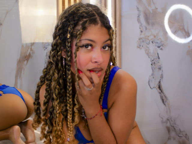 HaryAndKimmy with curly blonde braids, blue bra, red lipstick, and pink face paint, biting finger, looking at camera, ring light in background. Her ass is visible to the left.