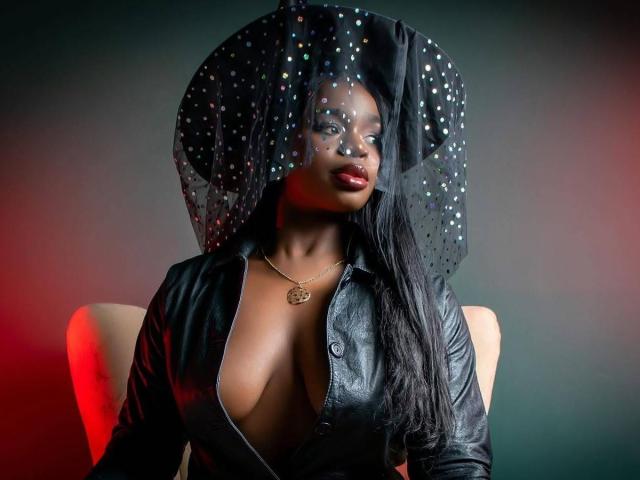 VenusWilly with dark skin, wearing a black leather jacket open to show her tits, a sparkling black headpiece, and a gold necklace, looking at the camera.