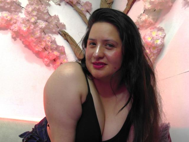 LauraCastillox with long black hair, pink lipstick, and black tank top, showing her big tits and smiling at the camera. Pink cherry blossom wall art in background.