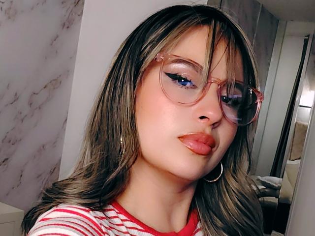 AmiiRosse met glanzende, roze lippen, blauwe oogschaduw en roze brillen, draagt een rood gestreepte shirt, oorringen en golvend bruin haar. Haar gezicht is lichtjes gedraaid, waardoor een schattige uitdrukking zichtbaar is..
