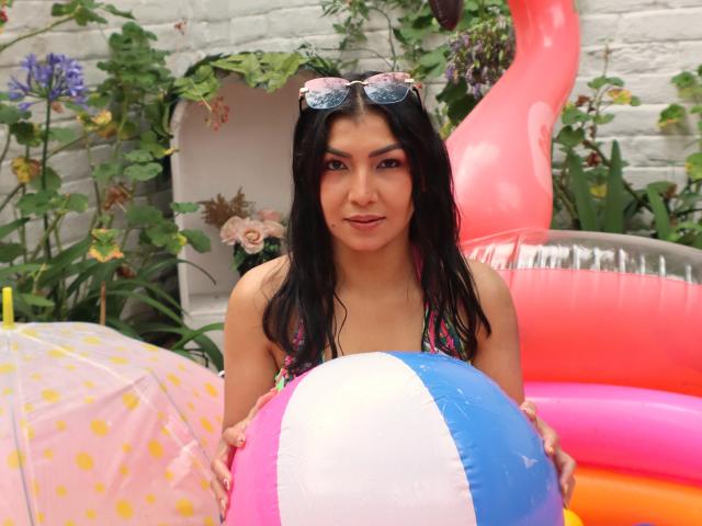ChanelBrouw, com cabelo preto molhado, usando um top de biquíni rosa, segurando uma bola de praia azul, branca e rosa, um brinquedo inflável rosa e um guarda-sol com bolinhas, com óculos de sol na cabeça..