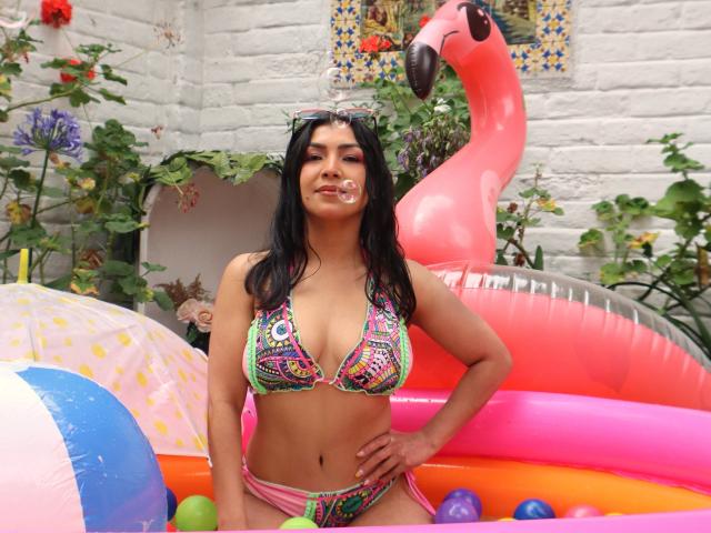 Chanel, vestida con un bikini colorido, mostrando sus pechos, con un flotador de flamenco rosa y una pelota de playa en el fondo, está de pie con una burbuja juguetona en la boca..