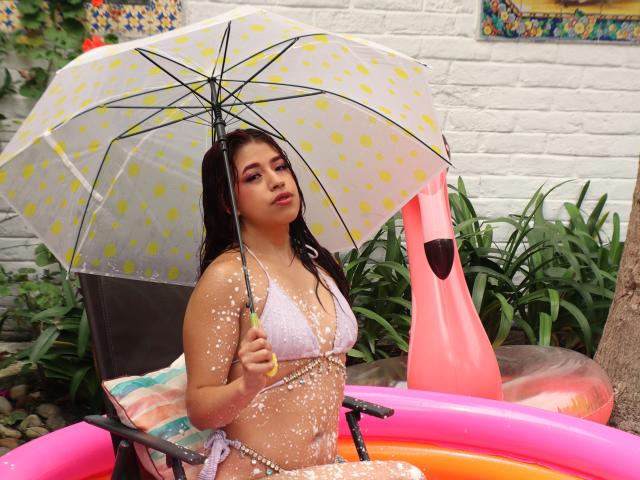 ElizaNior mit feuchten, gepunkteten Brüsten in einem rosa aufblasbaren Pool, die einen gelb gepunkteten Regenschirm hält, bedeckt mit weißen Wassertropfen, und ein weißes String-Bikini trägt..