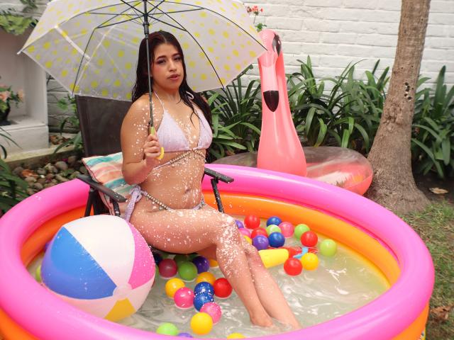 ElizaNior with wet brown hair, white bikini, holding polka-dot umbrella, sitting in pink inflatable pool with colorful balls and beach ball, water droplets on her tits and ass.