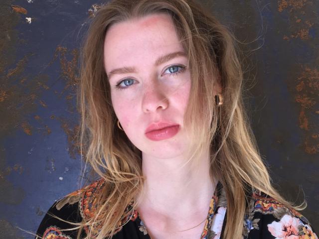 BettyDollores tiene la piel pálida, ojos azules y labios rosados, con cabello ondulado de color marrón claro, y lleva una camisa con estampado floral y pequeños aretes dorados, luciendo ligeramente fruncida..