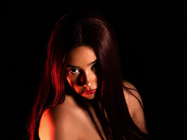 GraceBlue69 with long red hair, dark eyes, red lipstick, and bare shoulders, looking seductively at the camera in a dark, red-lit, studio portrait. Her tits are partially visible.