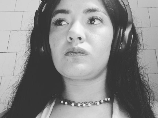 ArianithaXox with long dark hair, wearing headphones, looking up with a neutral expression, has a bead necklace, and a slightly chubby face with small moles on her chin.