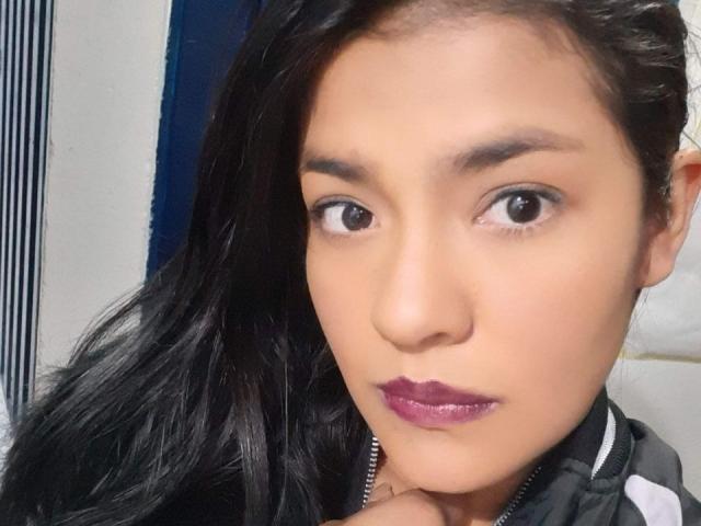 ArianithaXox has medium skin, dark brown eyes, and black hair. She's wearing purple lipstick, looking straight at the camera, with her hand near her chin.