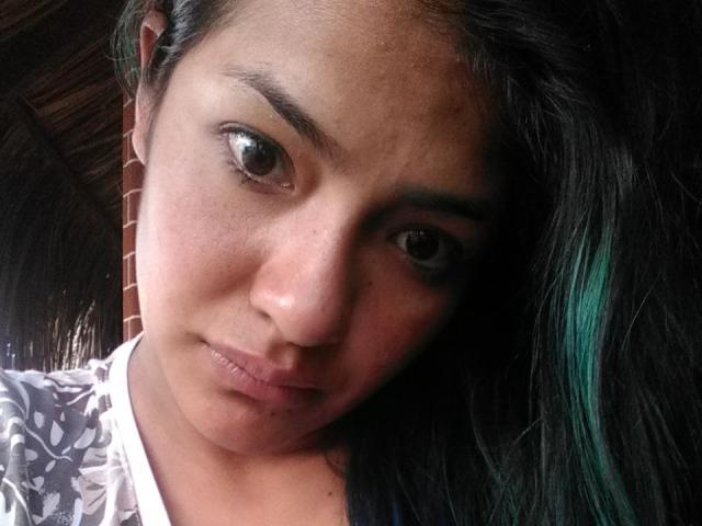 ArianithaXox's face in a selfie, showing brown eyes, slightly parted pink lips, dark hair with green highlights, and a white patterned shirt, leaning forward with a serious expression.