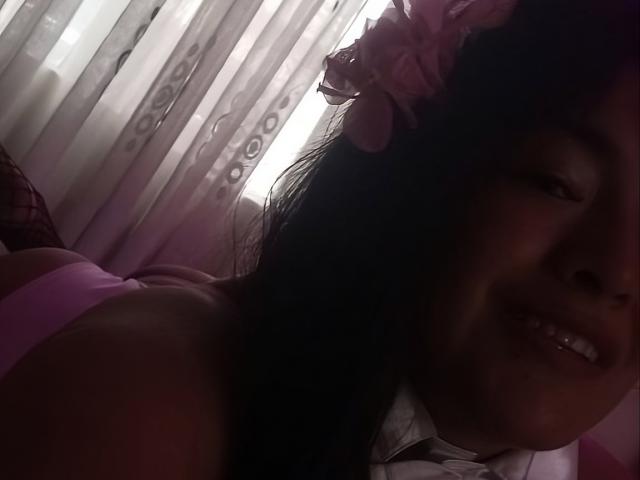 ArianithaXox with dark hair, pink flower in hair, smiling, wearing pink top, white collar, dimly lit, soft light, close-up, showing part of her tits and teeth.