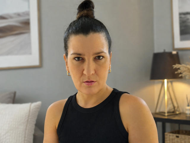 SilviaFreeya, avec ses cheveux noirs attachés en chignon, porte un haut noir sans manches, des boucles d'oreilles dorées et du rouge à lèvres rose, et regarde sérieusement l'appareil photo. Ses fesses ne sont pas visibles..