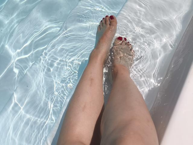 Dreia's benen in een zwembad, het water reikt tot aan haar tenen, rode nagellak, lichte huid, zonlicht dat rimpelt op het water, haar voeten staan iets uit elkaar, haar enkelbotten zijn zichtbaar..
