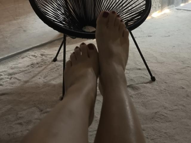 Dreia's bare feet with red-painted toes resting on a black, wire-framed chair, legs slightly crossed, showing smooth, fair skin. Shot from a lower angle, focusing on feet.