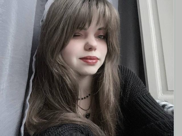 MiraRosse with winking eye, long brown hair, black knitted sweater, red lipstick, necklace, white window frame on right, gray curtain on left.