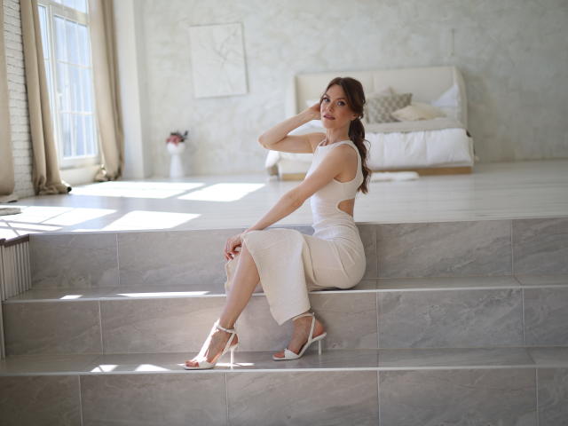 KatalinaJameson with tits in a white cutout dress, sitting on marble steps, hand on head, looking at camera, white heels, sunlight, bedroom in background.
