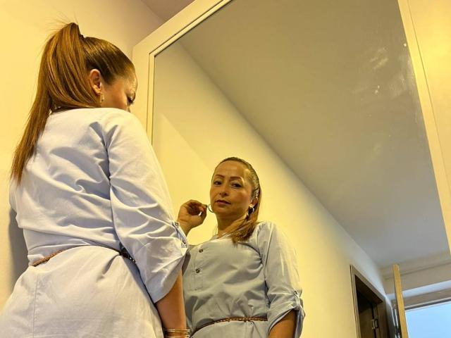 GianineLarose with brown ponytail, wearing light blue shirt, applying lipstick in front of mirror, reflection showing her focused face, white wall and door in background.