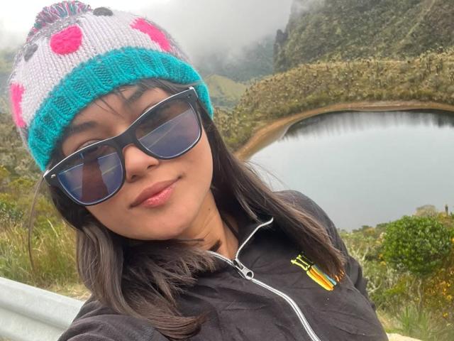 AshlyLee with dark brown hair, wearing a turquoise and pink knit beanie, black sunglasses, and a black zip-up jacket, smiling near a misty, mountainous lake.