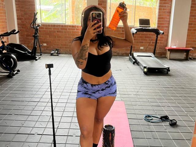 AlanaLuxe with black top, purple tie-dye shorts, holding orange water bottle, reflecting in mirror, gym with treadmill, bike, and weights, showing tattooed arm and large tits.