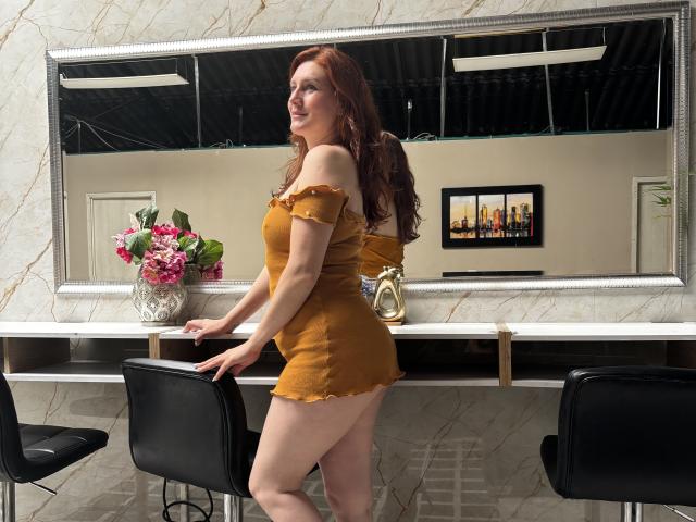 KatyaRogersX, red-haired woman in a tight, off-shoulder, yellow dress, showing her ass, leaning on a white marble bar, with black chairs, and a flower vase.
