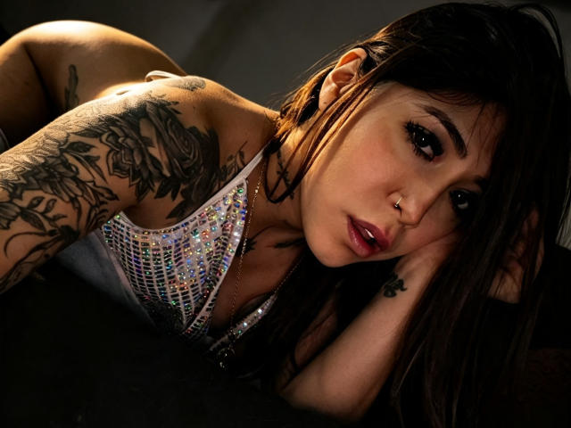 Sthephysexy is lying down, looking at the camera with dark, shiny eyes. She has tattoos on her arms, a nose ring, and is wearing a sparkly, mesh top.