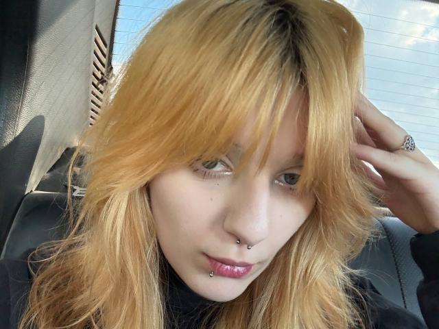 ForenCostler has blonde hair, green eyes, and multiple facial piercings including a nose ring and lower lip ring. Sitting in a car, hand in hair, looking at camera.