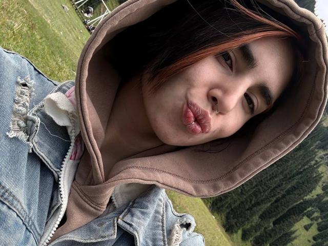 TameikaMacclellan with pouty lips, brown hoodie, and blue jeans, showing off her nose ring and dark red hair, in a grassy field with trees in the background.