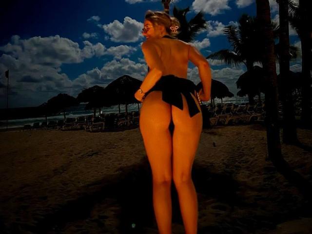 MillieHartwell stands topless on a beach at night, black thong, back to camera, hands on butt, palm trees, dark blue sky with clouds, sand, and ocean in background.
