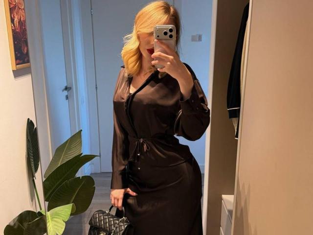 MillieHartwell with blonde hair, wearing a black satin dress, holding a phone in front of her face, showing her tits and ass, standing in a room with a plant.