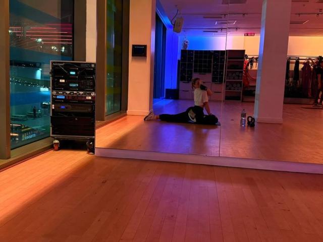 MillieHartwell, lying on wooden floor in front of mirror, doing a back stretch with hands on floor, wearing black leggings and white shirt, water bottle nearby.