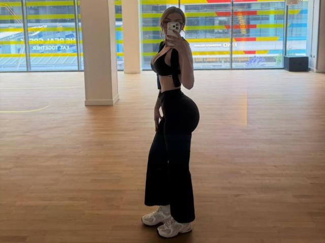MillieHartwell with big ass, small waist, wearing black sports bra, high-waisted black pants, white sneakers, taking mirror selfie in wooden floor gym, holding phone, covering face.