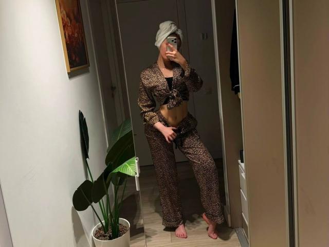 MillieHartwell with blonde hair, wearing leopard print crop top and pants, holding phone, covering pussy, barefoot, standing in mirror, potted plant on left.