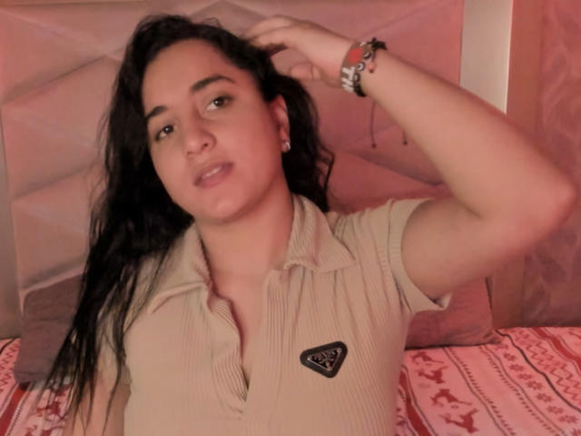 LisaStarkm with dark hair, wearing a beige ribbed shirt, hand in hair, brown eyes, pinkish skin, earrings, bracelet, lying on red-patterned bed, slightly open mouth, relaxed expression.