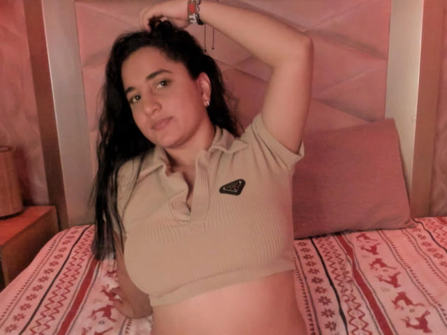 LisaStarkm with dark hair, beige ribbed crop top, small black logo, lifting arm, showing midriff, sitting on bed with pink patterned sheets, looking at camera, slight smile.