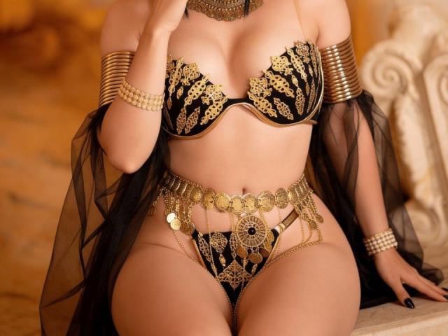 ElzaKomx in a black and gold lace bra and matching panties with a chain belt, wearing gold bangles and a sheer black cape, showing off her tits and pussy.
