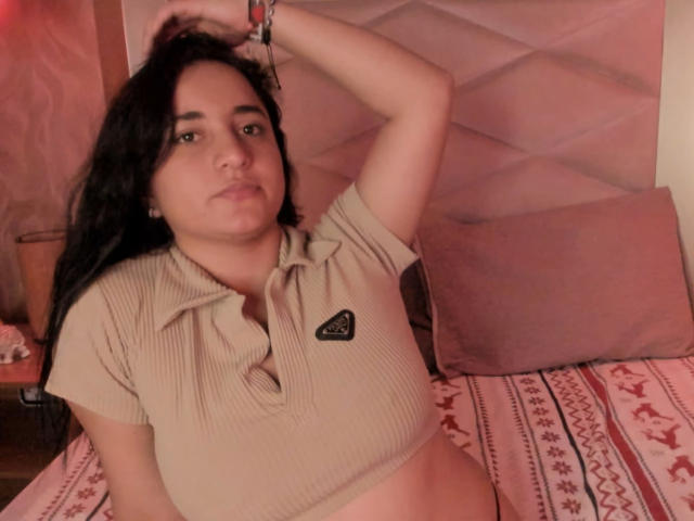 LisaStarkm with long dark hair, wearing a beige ribbed crop top, exposing her belly, and her hand in her hair, sitting on a bed with pink pillows.