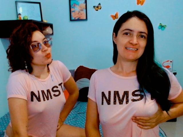 LunaVenus with curly red hair and glasses, wearing a pink NMST shirt, sitting with hands on knees. LunaVenus with straight black hair, holding her tits, smiling.