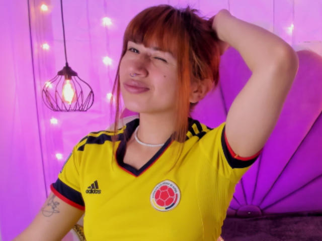 NaraStorn with red hair, winking, wearing a yellow Adidas soccer shirt, black and red trim, and a pink nose ring, hand in hair, smiling, purple light.