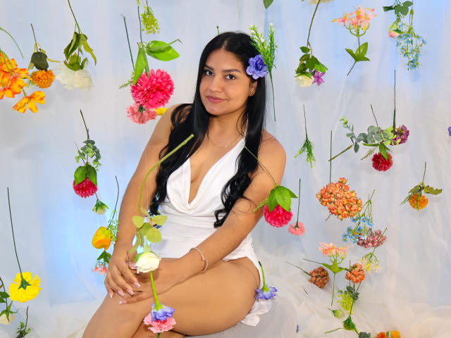 AlmaRoses, con cabello negro largo, vestida con un vestido blanco, sentada entre flores coloridas, tiene piel morena, senos pequeños y una flor púrpura en el cabello, sonriendo suavemente..