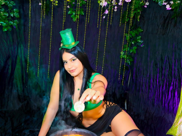 AlmaRoses com um chapéu verde, cabelo preto comprido, sutiã verde, shorts pretos e colar verde, segurando uma moeda, estendendo o braço, iluminação roxa e verde, sorrindo, fundo preto..