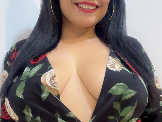 CharlotteCrofort has large, creamy-boob tits with a deep V-neck floral dress, red lipstick, and gold hoop earrings, smiling with a nose ring. Her black hair is long.