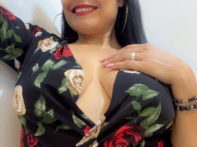 CharlotteCrofort with large boobs in a black floral dress, red and beige roses, gold hoop earrings, red lipstick, ring on right hand, and pink nail polish, smiling.