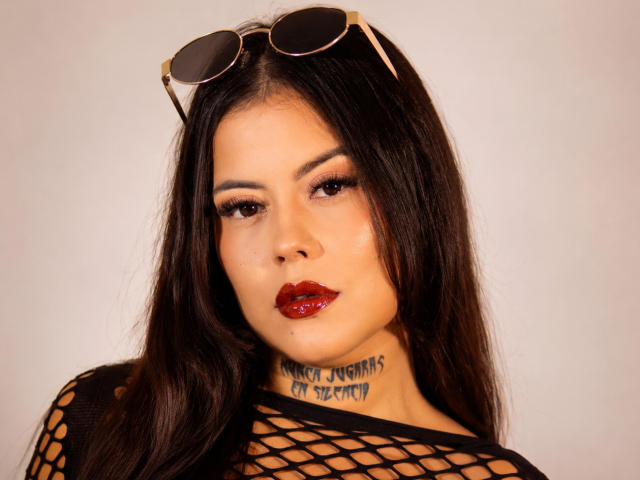 ElectraRoss with long black hair, red lipstick, and fishnet top, wearing sunglasses on her head, has a tattoo under her chin, and a serious, slightly seductive expression.