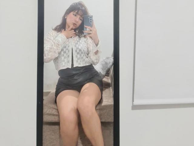 HairyValeri with medium tits, wearing a checkered blouse and black skirt, sitting on a bench, taking a mirror selfie with her phone, legs crossed, in a white room.