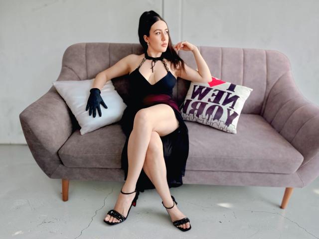MabyLegrand sits on a gray sofa, wearing a black halter dress, black gloves, and strappy heels, with her legs crossed, showing her ass, and a relaxed, confident expression.