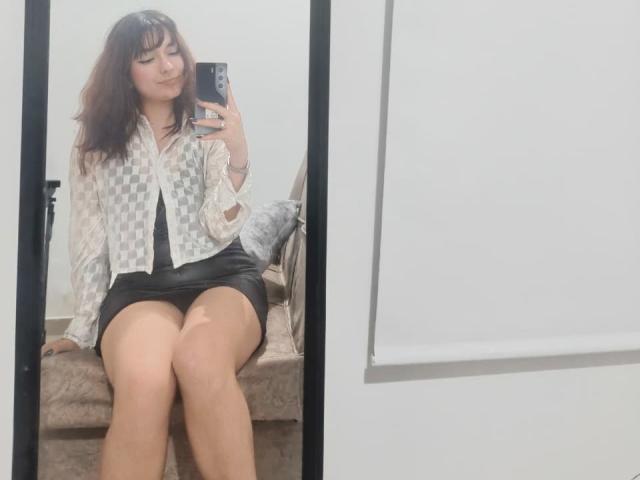 HairyValeri with brown hair, wearing a checkered shirt and black skirt, sitting on a beige couch, holding a phone, showing off her thighs and smiling in a mirror.